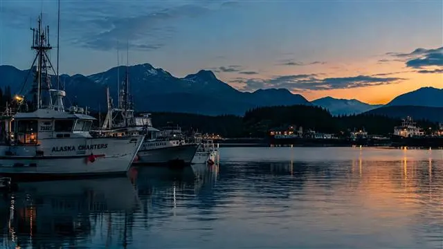 Calm Alaskan coastal harbor at dusk with moored charter boats and forested mountains Calm Alaskan coastal harbor at dusk with moored charter boats and forested mountains